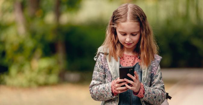 Pretty school girl with smartphone outdoors. Cute female kid standing with cell phone in autumn park