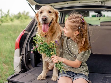 Beautiful little girl sitting with golden retriever dog in the car trunk holding flower bouquet and hugging pet. Child kid with purebred doggy and summer plants in the vehicle at the nature