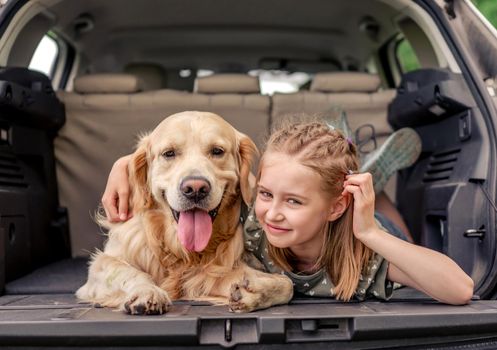 Beautiful little girl lying with golden retriever dog in the car trunk and looking back. Child kid with purebred doggy pet with tonque out in the vehicle at the nature together