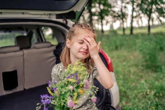 Beautiful little girl with cute hairstyle sitting in the car trunk holding flower bouquet and close her eyes with her hand. Child kid with summer plants in the vehicle at the nature