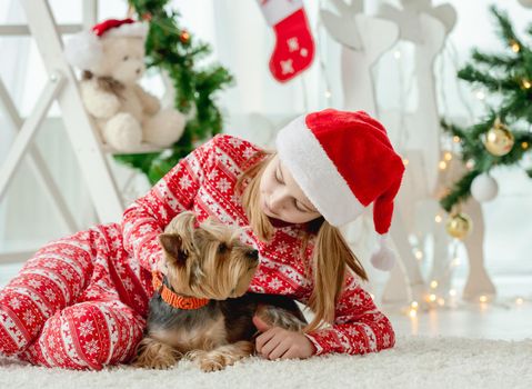 Child girl with dog sitting on floor with Christmas tree on background, looking at camera and smiling. Kid and pet doggy enjoying New Year time and decoration at home