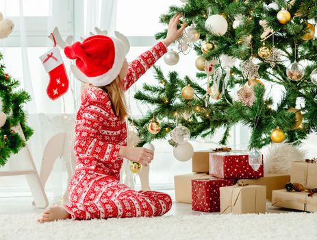 Child girl wearing red Santa hat decorated Christmas tree. Kid celebrating New Year with gifts