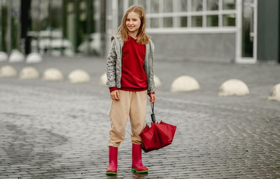 Preteen girl wearing roober boots and holding umbrella outdoors in rainy autumn day. Pretty child in gumboots portrait at street