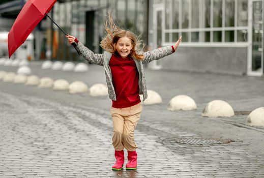 Preteen girl wearing roober boots and holding umbrella outdoors in rainy autumn day smiling. Pretty child in gumboots having fun at wet street