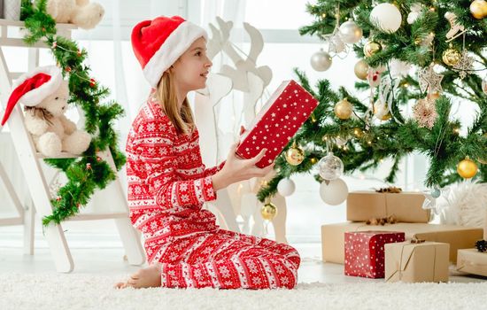 Child girl holding in Christmas time red gift box at home. Kid celebrating New Year with presents and Christmas tree