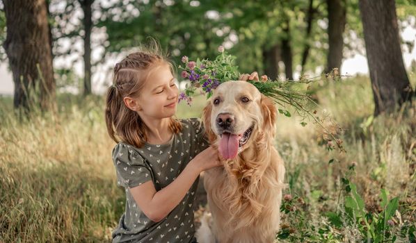 Portrait of beautiful preteen girl petting golden retriever dog and looking at him outdoors. Kid with doggy pet in the field in summer time