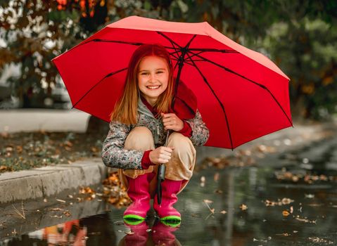 Preteen girl wearing rubber boots sitting under umbrella at autumn at street in rainy day. Pretty female kid in gumboots outdoors