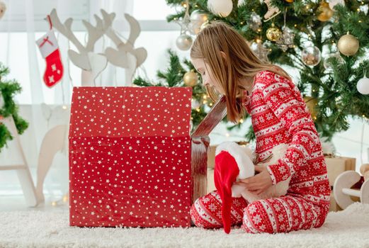 Pretty child girl open Christmas red gift box at home with traditional decorated tree. Kid celebrating New Year with presents