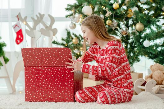 Pretty child girl open Christmas red gift box at home with traditional decorated tree. Kid celebrating New Year with presents