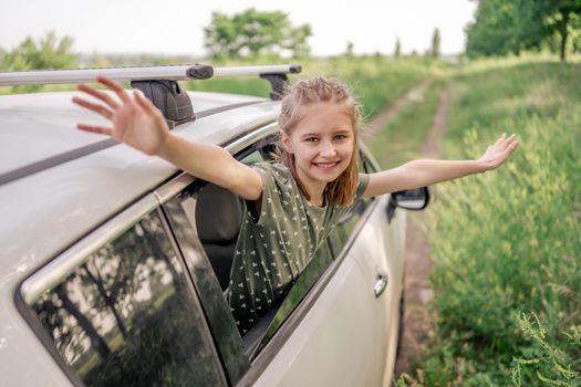 Beautiful preteen girl sitting in the car and looking out the window open, smiling and holding hands up in the field. Happy child kid in the vehicle outdoors during summer journey