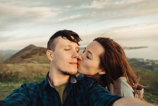 Traveler young loving couple taking self-portrait on peak of mountain. Woman kissing a man with closed eyes