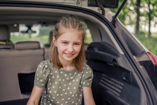 Closeup portrait of cute preteen girl with beautiful hairstyle sitting in the car trunk and smiling looking at the camera. Happy child kid in the vehicle at the nature during summer trip