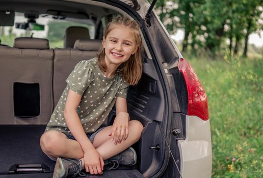 Preteen girl with beautiful hairstyle sitting in the car trunk and smiling looking at the camera. Happy child kid in the vehicle at the nature during summer trip