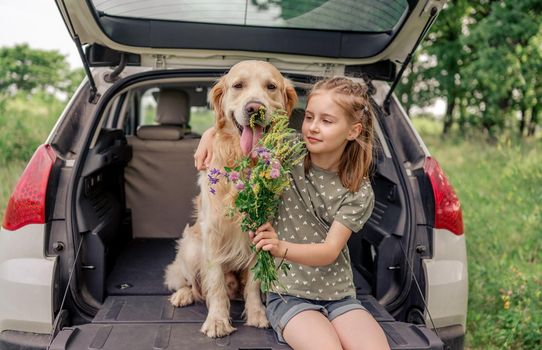 Beautiful little girl sitting with golden retriever dog in the car trunk, hugging it and holding flower bouquet. Child kid with purebred doggy pet and summer plants in the vehicle at the nature