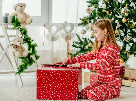 Child girl unpacks Christmas red gift box at home. Kid celebrating New Year with presents