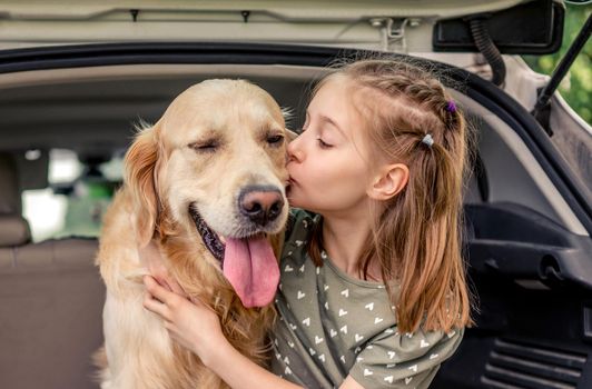 Beautiful little girl sitting with golden retriever dog in the car trunk, hugging pet and kisses it. Child kid with purebred doggy in the vehicle at the nature