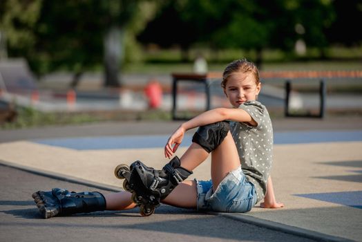 Beautiful preteen girl wearing roller skates sitting on ground and looking at camera