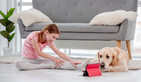 Little girl sitting on the floor with golden retriever dog and stretch her legs during online workout with tablet