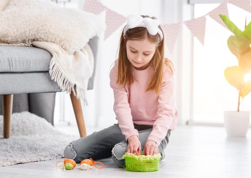 Little girl sitting on the floor with decorated basket and painted eggs preparing for Easter day