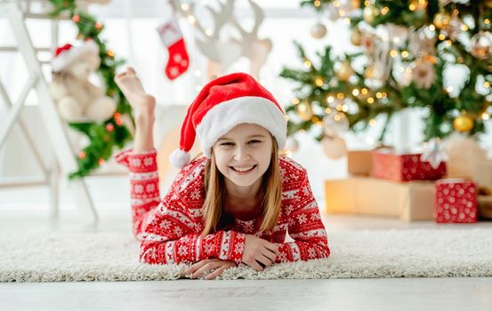 Pretty child girl lying on floor at Christmas home with traditional decorated tree. Happy smiling kid in New Year time