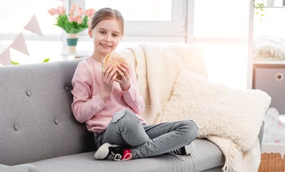 Little smiling girl sitting on the sofa at home and holding hamburger at her hands