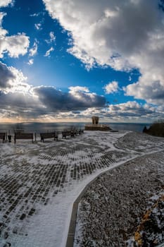 Panoramic wide angle view on snow-covered park on the embankment against the background of the winter blue sea. Promenade with a beautiful view of the sea on a cloudy day