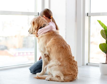 Little girl sitting on the floor next to panoramic window with golden retriever dog and hugging him