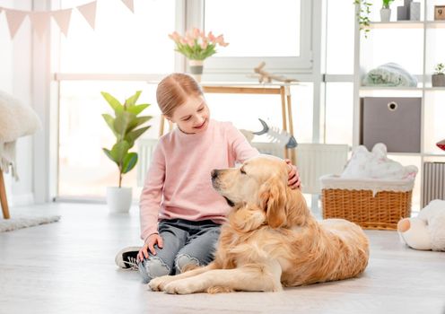 Little girl sitting in the sunny room on the floor with golden retriever dog and smiling