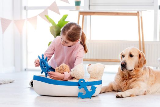 Little girl sitting on the floor and playing with sea ship with golden retriever dog