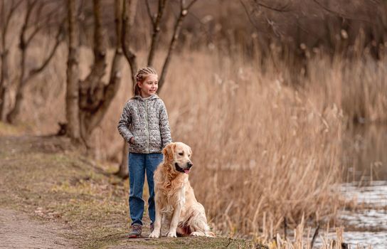 Preteen beautiful girl standing with golden retriever dog on the nature and looking at the river