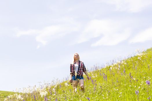 Happy adventurer female stands on the green mountain slope among flowering pink rhododendrons and looking into the distance. Epic travel in the mountains. Wide angle.