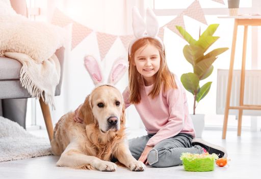 Little girl with golden retriever dog wearing bunny ears sitting on the floor and looking at the camera in room decorated for Easter