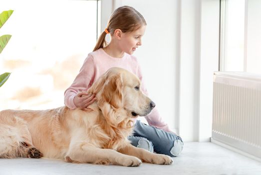 Little girl and golden retriever dog sitting together on the floor and looking out the window