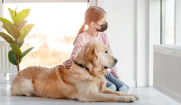 Little girl wearing black mask and golden retriever dog sitting together on the floor and looking out the window