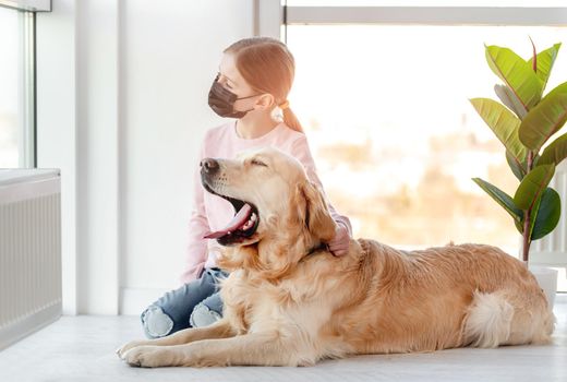 Little girl in black mask sitting with yawning golden retriever dog and looking out the window