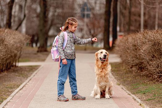 Little beautiful girl with backpack training golden retriever dog on the nature