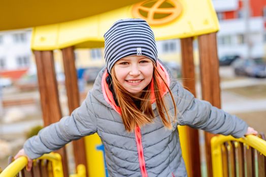 Little smiling girl wearing coat and hat standing on playground and looking at the camera