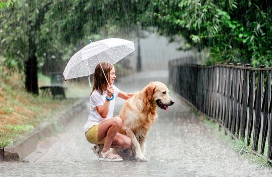 Girl with golden retriever dog during rain sitting under umbrella outside. Preteen kid with doggy pet in rainy day
