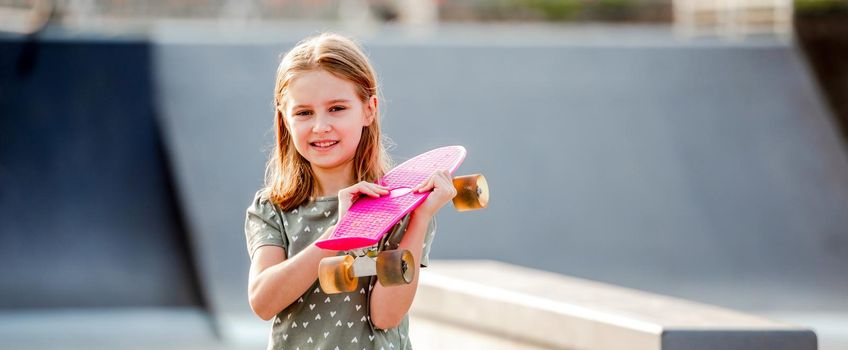 Sweet girl holding skateboard and looking at camera outdoors. Female skater child with skate in the city