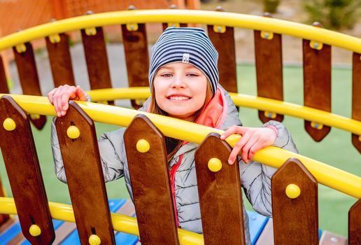 Little girl sitting behind wooden fence with yellow metal handrail on playground and smiling