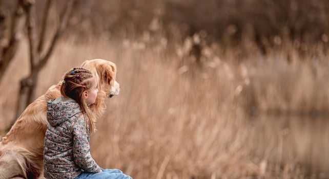 Preteen girl sitting with golden retriever dog close to river in spring time