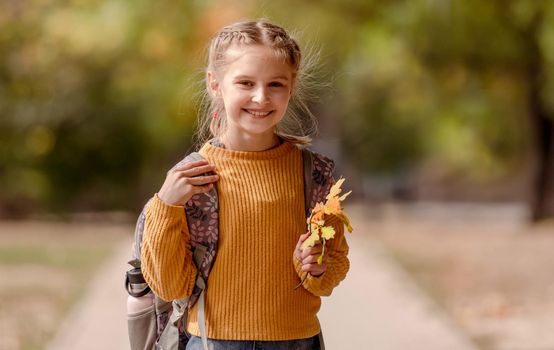 Preteen school girl with backpack holding yellow leaves in her hand, looking at camera and smiling. Pretty pupil female kid after education class at autumn