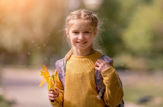 Preteen school girl with backpack holding yellow leaves in her hand, looking at camera and smiling. Pretty pupil female kid after education class at autumn