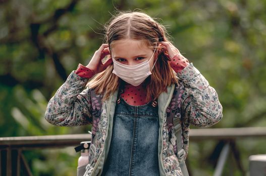 Pretty school girl wearing face mask and looking at camera. Beautiful female kid after school in pandemic coronavirus time