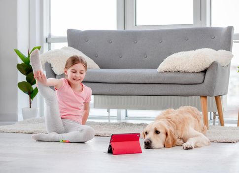 Child yoga online exercises with tablet in coronavirus time. Golden retriever dog with girl at home