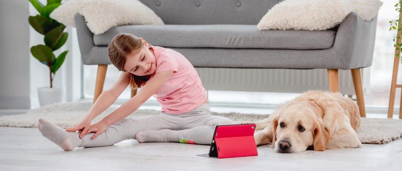 Child girl making yoga online workout at home with tablet while golden retriever dog laying close to her on the floor