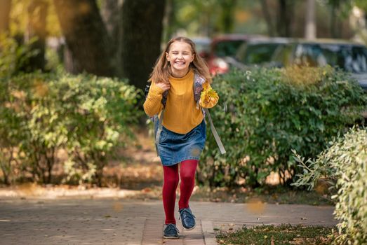 Preteen school girl with backpack holding yellow leaves in her hand, running and smiling. Pretty pupil female kid having fun in autumn park after lessons
