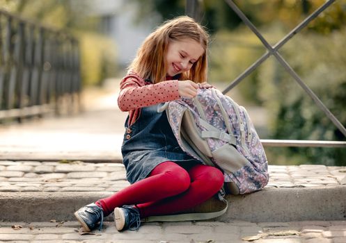 Preteen school girl looking in backpack, sitting on ground and smiling. Sweet pupil female kid resting after education class in autumn park