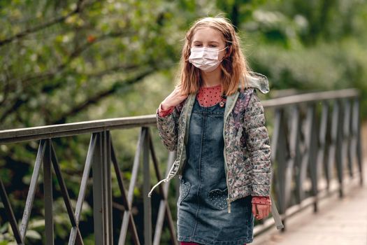Pretty school girl wearing face mask standing in autumn park and looking at camera. Beautiful female kid after school in pandemic coronavirus covid time