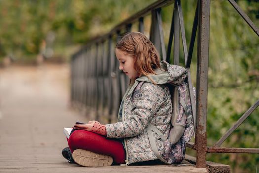 Preteen school girl with backpack sitting on ground and reading book outdoors. Sweet pupil female kid studying in autumn park